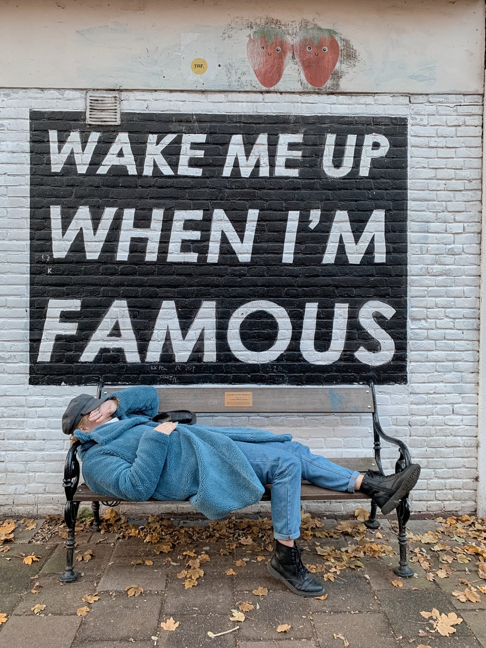 Free Side view of anonymous lady in fashionable coat and jeans covering face with hand while lying on bench placed on street near Wake Me Up When I M Famous inscription Stock Photo